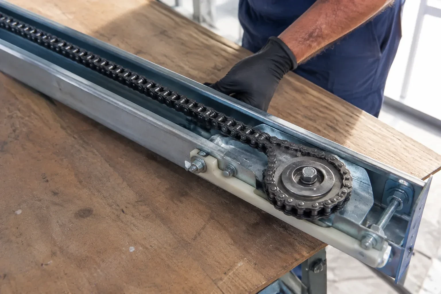 Gloved hand resting on a steel opener rail laid on a wooden bench, showing a drive chain looped around a sprocket wheel. — Chain-Drive Opener Repair, Scotch Block