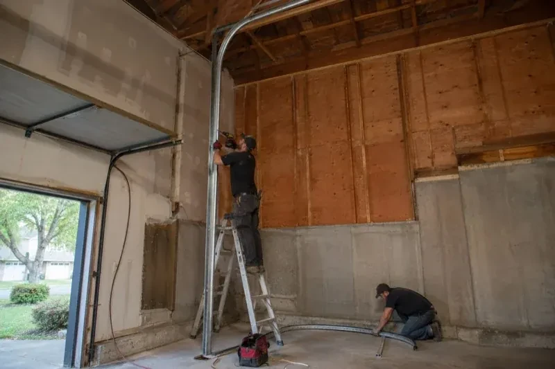 One person on a stepladder working beside a tall curved garage-door track while another kneels beside loose track sections in an unfinished garage. — High-Lift Garage Door Conversion, Halton Hills